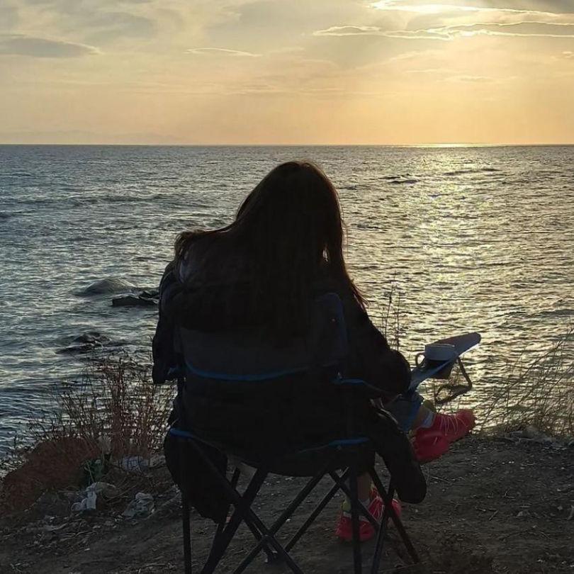 The silhouette of a girl sitting on a camping chair in front of the setting sun over a lake.
