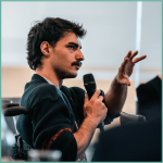 A man with an excellent moustache holding a microphone and gesturing towards the audience.