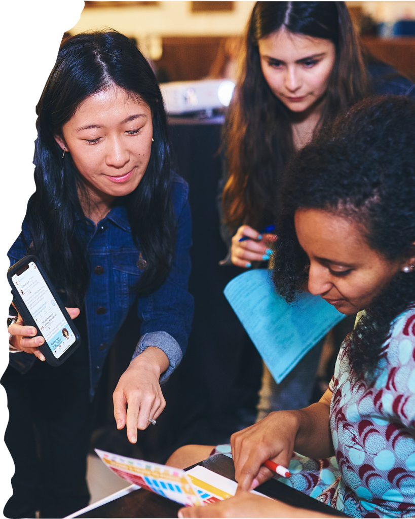 Three women are leaning over a paper, with one woman showing her phone and pointing at the paper.